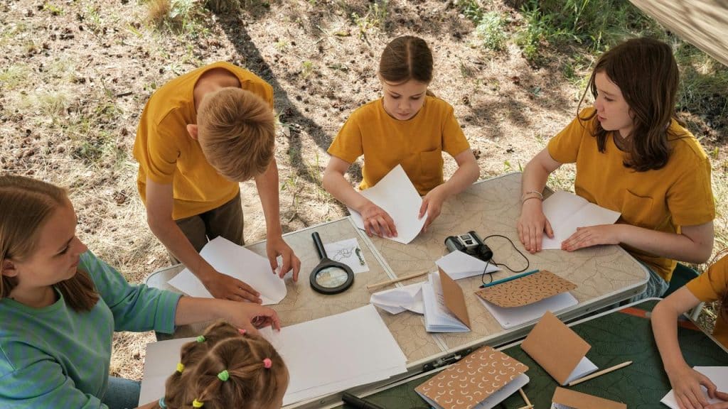 A group of kids folding paper at an outdoor table.