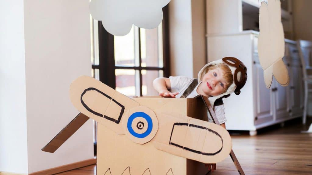 A child playing in a cardboard airplane wearing aviator gear.