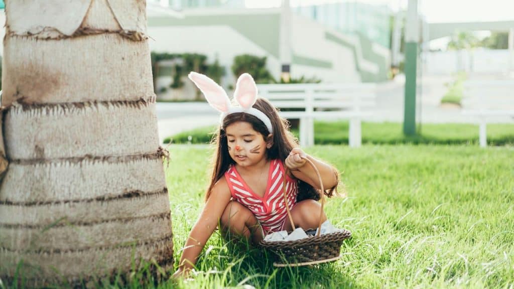 A girl in bunny ears collecting eggs with a basket on grass.