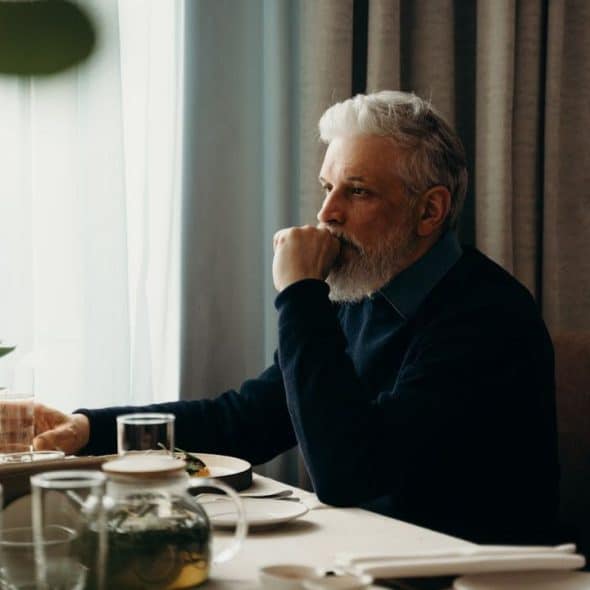 A man with gray hair and a beard sits at a table and looks out the window.