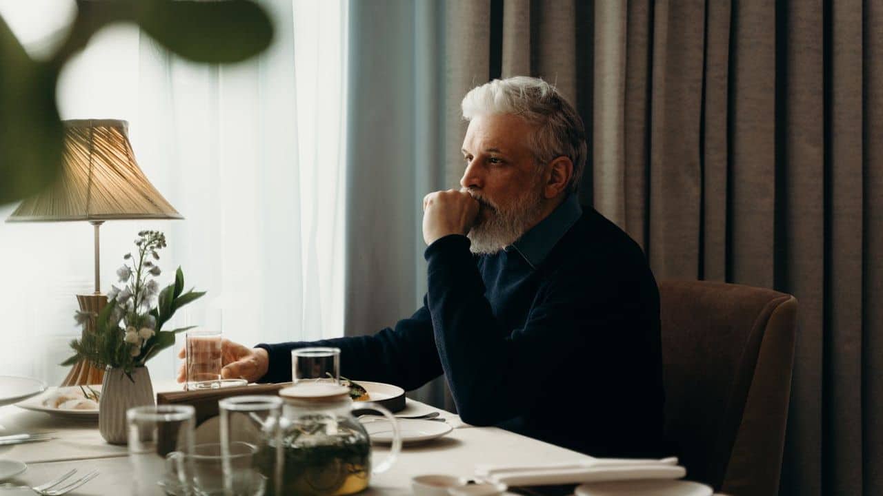 A man with gray hair and a beard sits at a table and looks out the window.