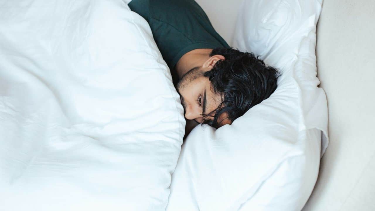 A man is sleeping in bed with his face mostly covered by a pillow and comforter.