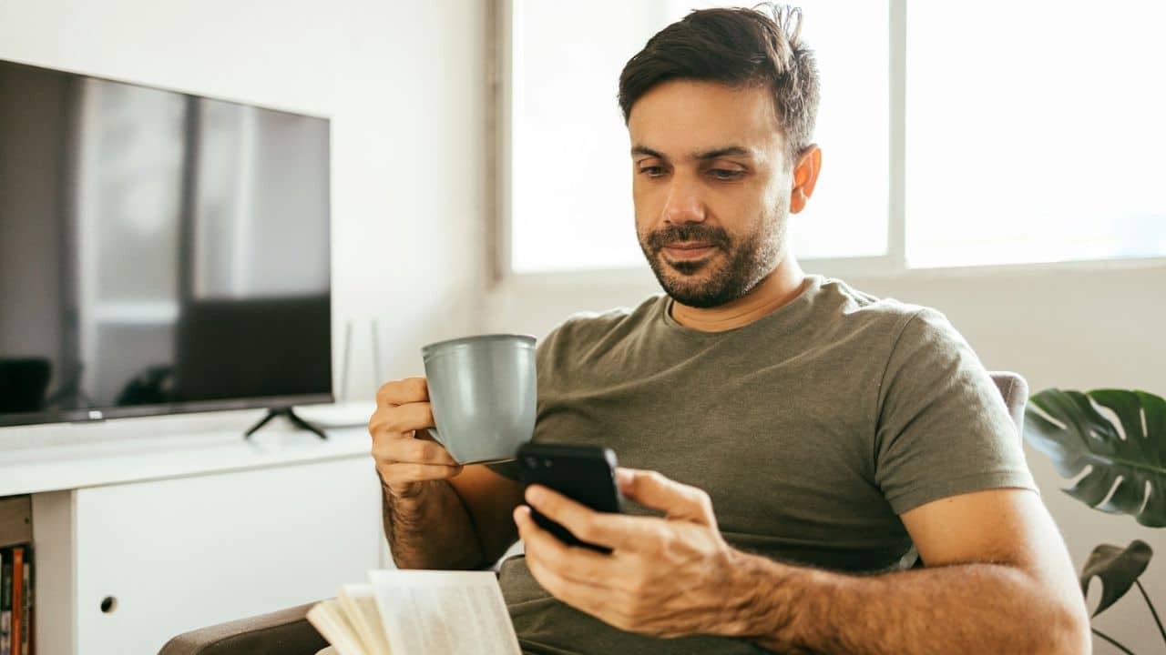 A man is sitting on a chair, drinking from a mug and looking at his phone.