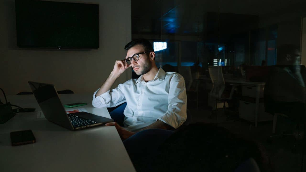 A man wearing glasses and a white shirt works late at his laptop in an office.
