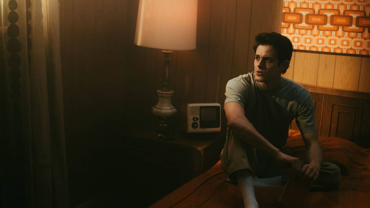 A man sits on a bed with a lamp and radio nearby, looking away from the camera.