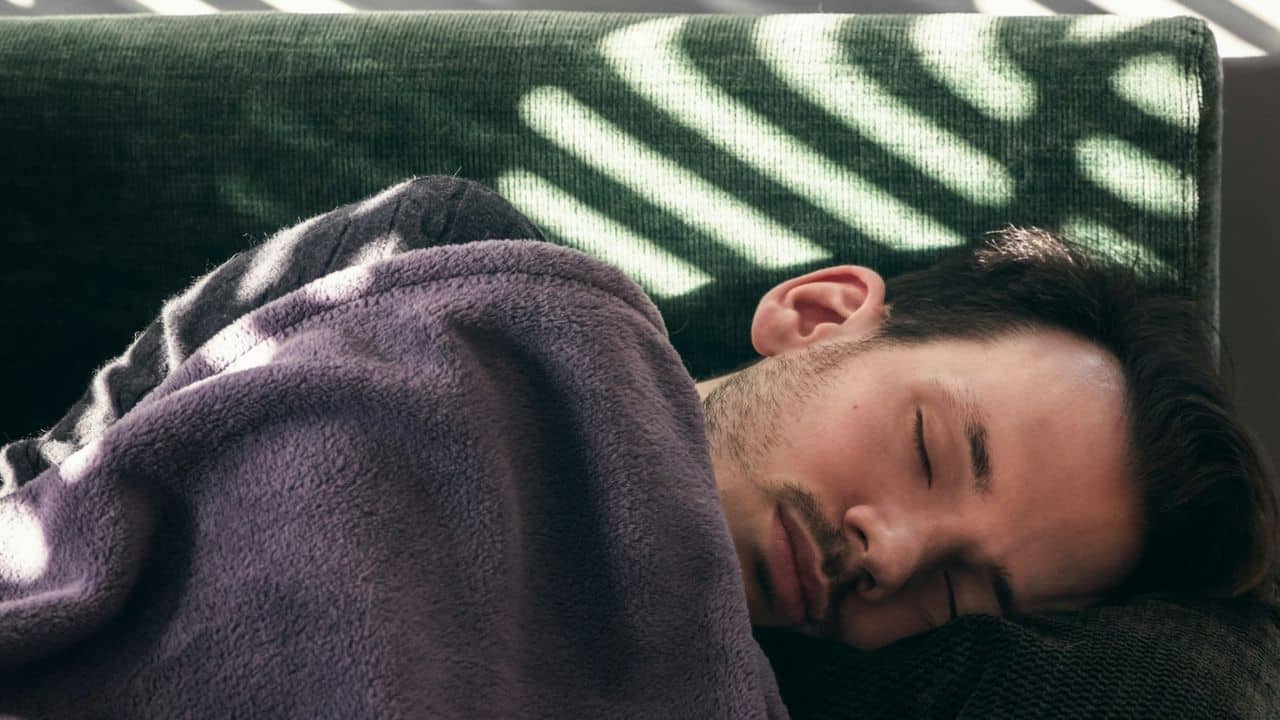 A man is sleeping on a couch covered in a blanket with sunlight shining on him.