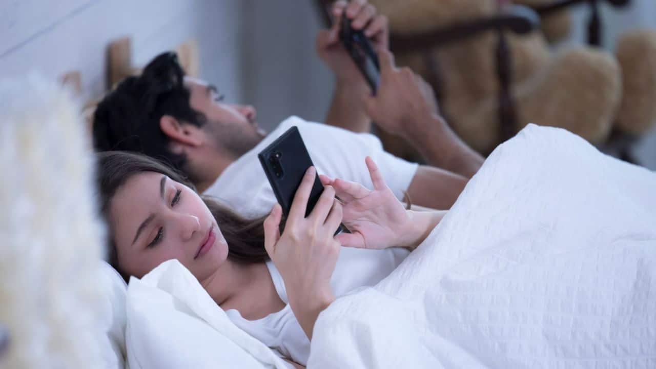 A man and woman lie in bed, each looking at their own phone.
