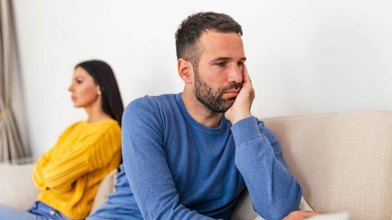 A man and a woman sit on a couch with their backs to each other. The man looks away with a hand on his face.