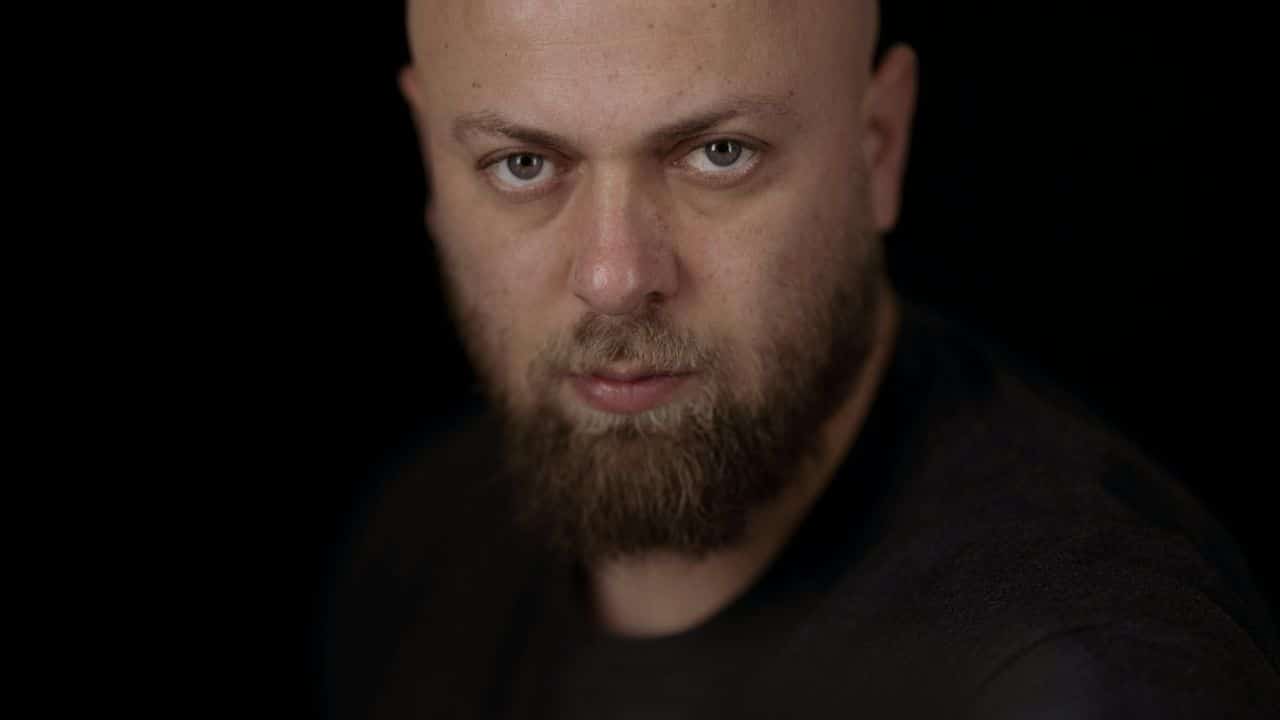 A close-up, head-on shot of a bald man with a beard staring at the camera.
