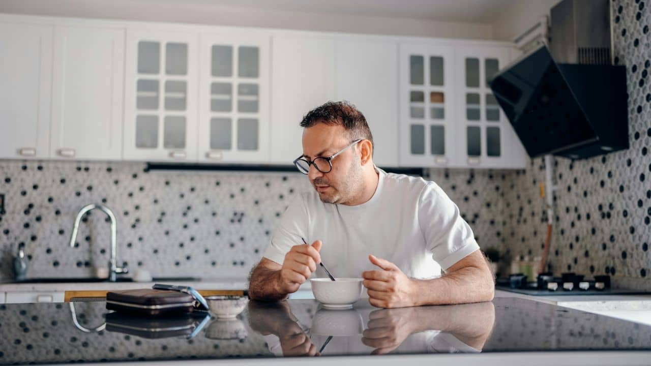 A man wearing glasses and a white shirt sits at a kitchen counter with a bowl and tablet.