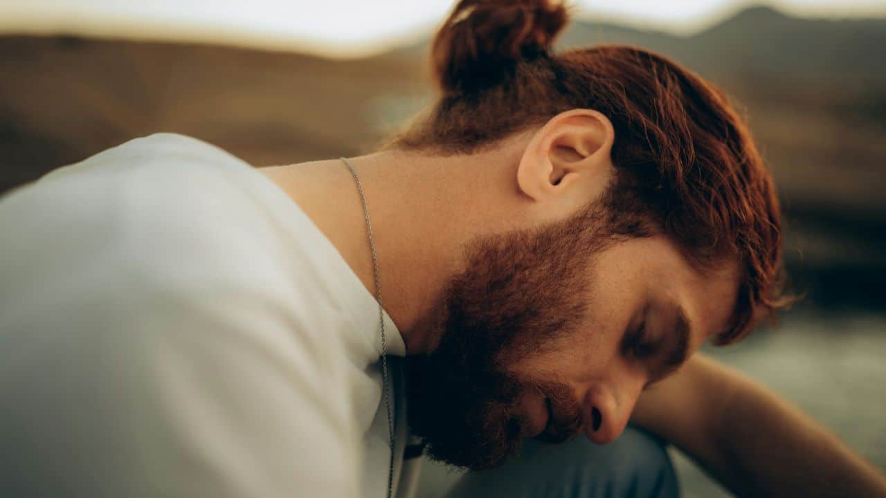 A man with red hair and a beard sits outdoors with his eyes closed.