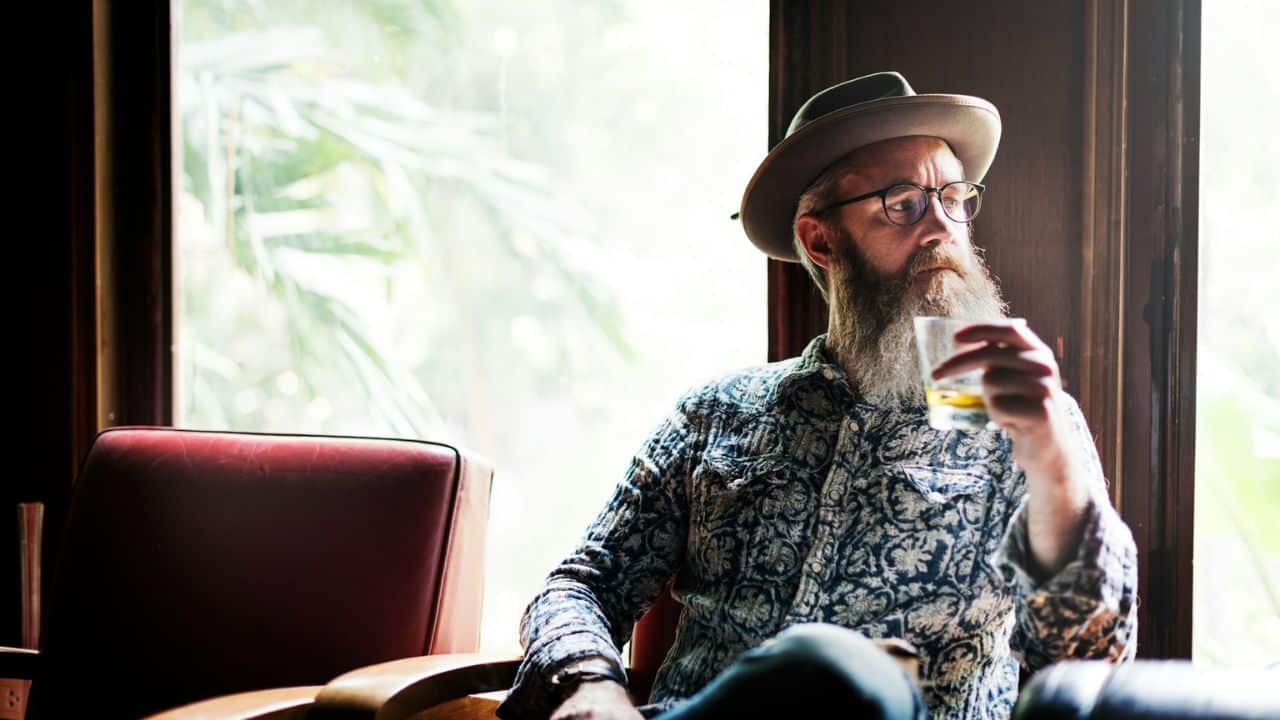 A man with a long beard and a fedora sits in a chair, holding a glass.
