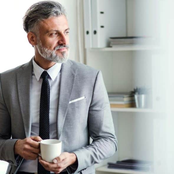 A handsome, gray-haired man in a suit holds a mug and looks away.