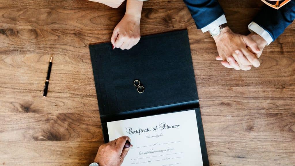 A high-angle view of two people signing a divorce certificate, with wedding rings on top.