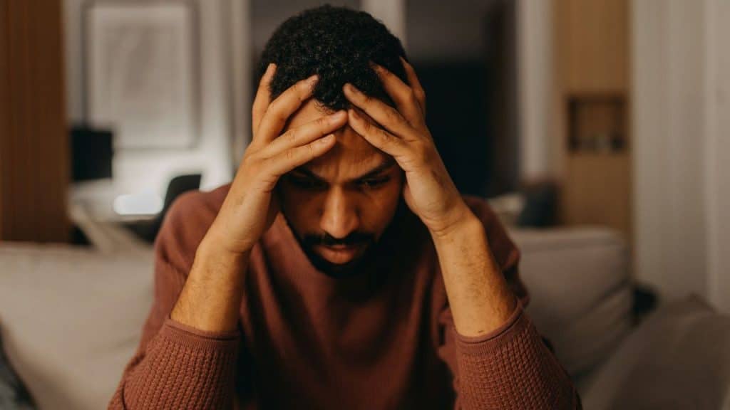 A stressed man sits with his hands on his head, looking down.