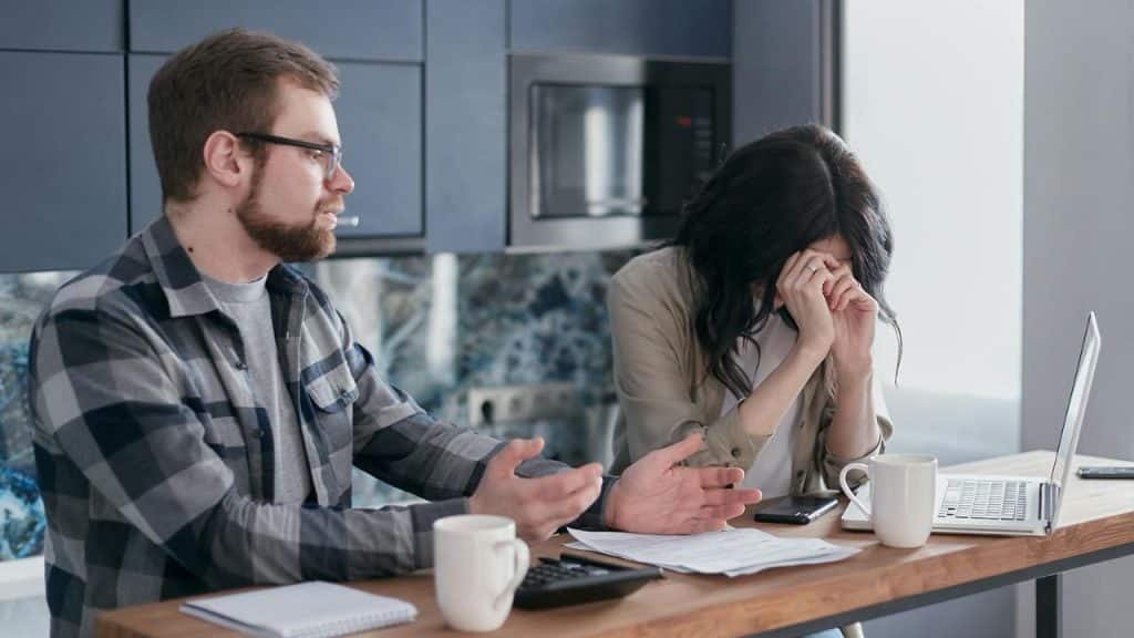 A frustrated man talks to a woman covering her face with her hands, sitting at a table with papers.