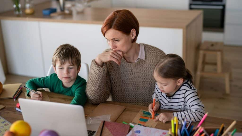 A stressed mother with a hand on her face looks at her children coloring.