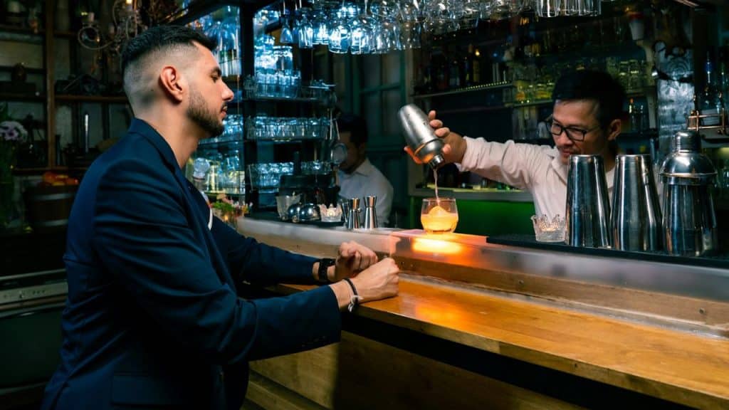 A bartender pours a drink for a man sitting alone at a bar.