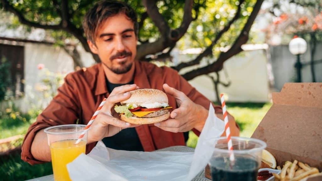 A man holds a hamburger with both hands, sitting at a table outdoors.
