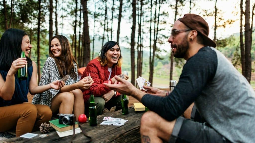 A diverse group of young adults laughs while playing cards and drinking beer in the woods.