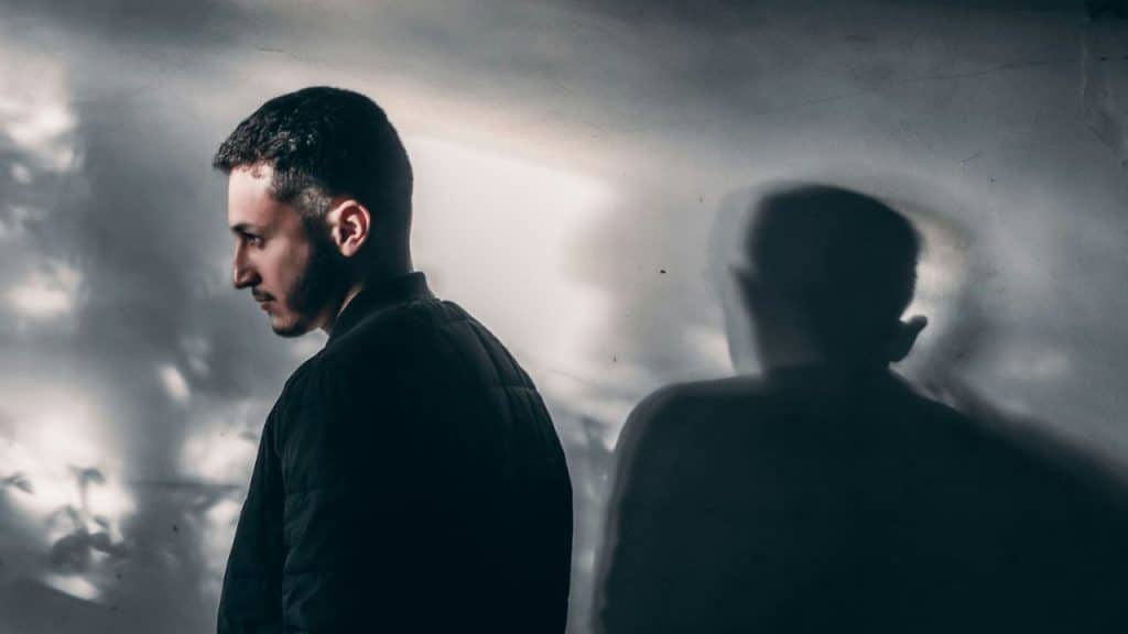 A somber-looking man in a black jacket stands, with a shadow of a person behind him.