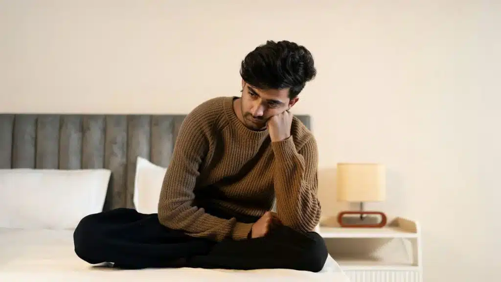 A sullen man in a brown sweater and black pants sits on a bed.