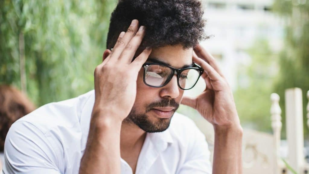 A distressed man in glasses holds his head in his hands, looking down.