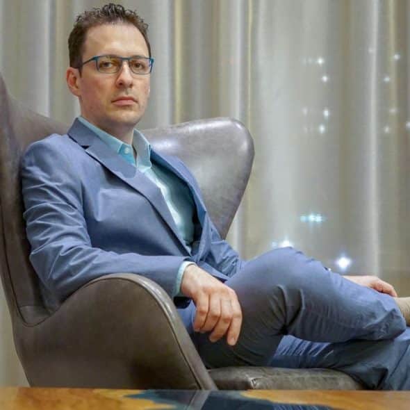 A serious man in a light blue suit and glasses sits in a brown leather armchair.