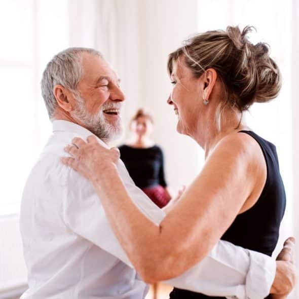 A cheerful older couple in a bright room embraces, seemingly dancing, with a blurry figure in the background.