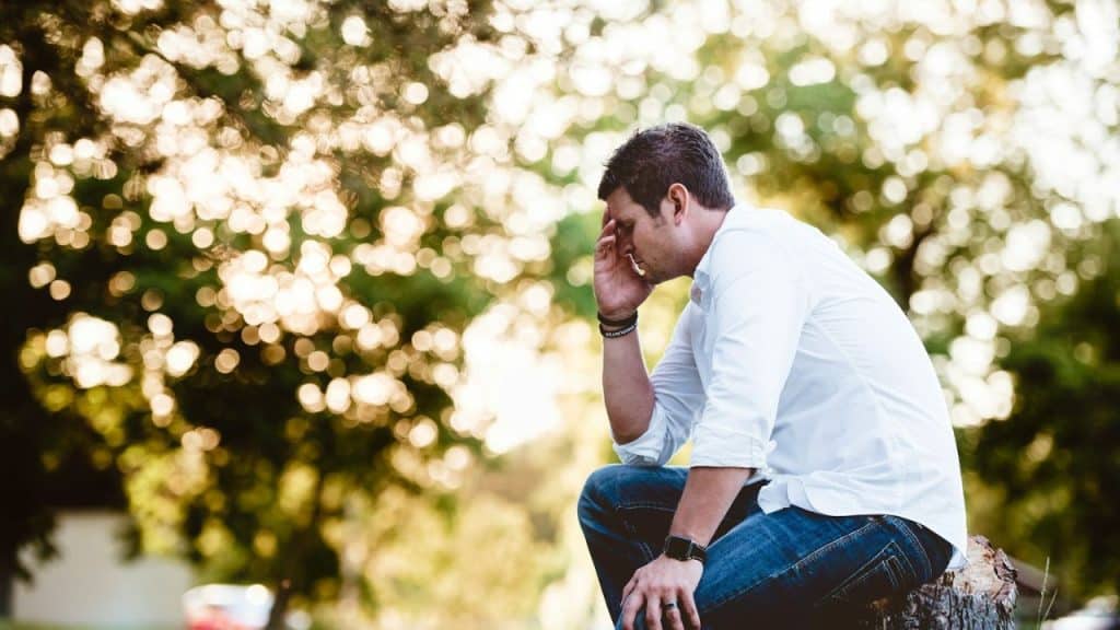 A man in a white shirt and jeans sits on a tree stump, holding his head.