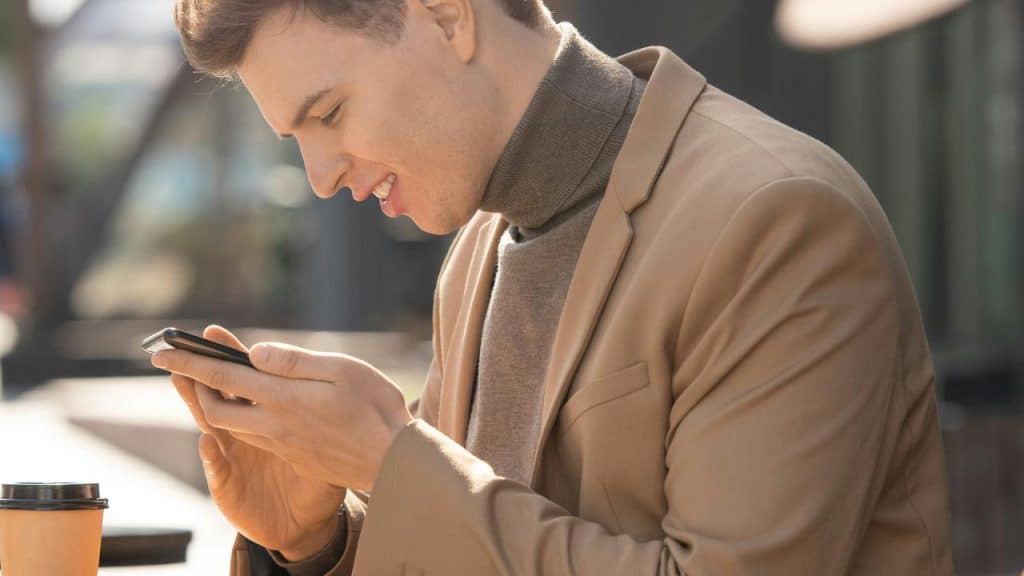 A man in a brown coat smiling while looking at his phone.