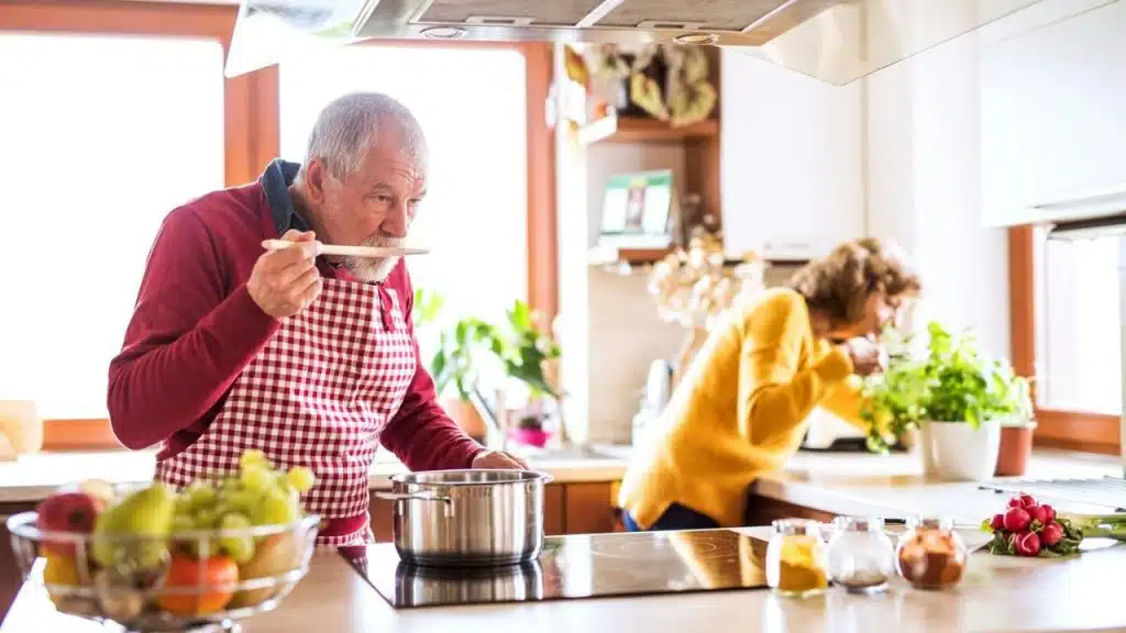 A senior couple is in a kitchen, with the man in the foreground tasting food and the woman smelling herbs.