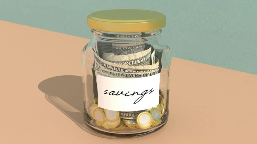 A glass jar labeled "savings" contains dollar bills and coins on a two-toned background.