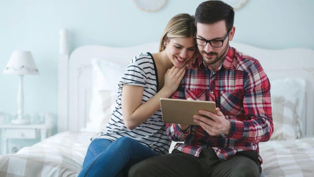A smiling couple sits on a bed, looking at a tablet together.