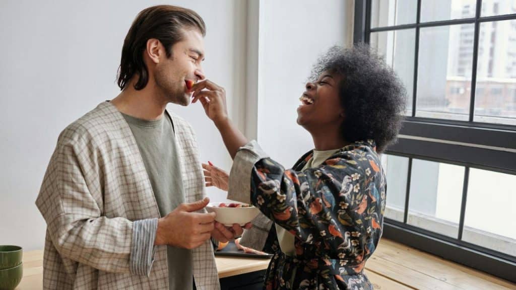 A joyous woman with an afro feeds a smiling man a strawberry in a sunlit room.