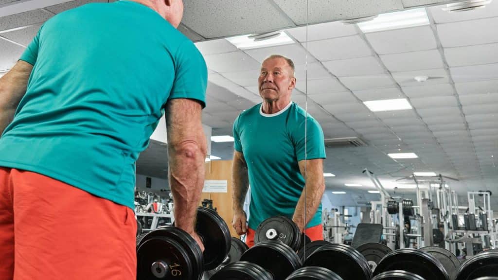An older man lifting dumbbells while looking in the mirror at the gym.