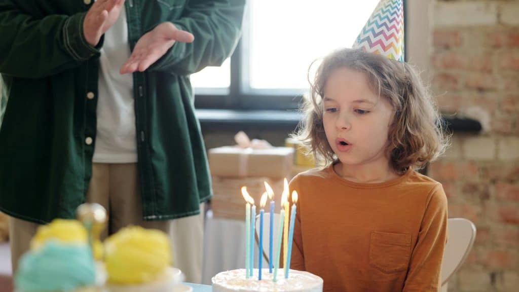 A kid wearing a party hat blowing out candles on a birthday cake.