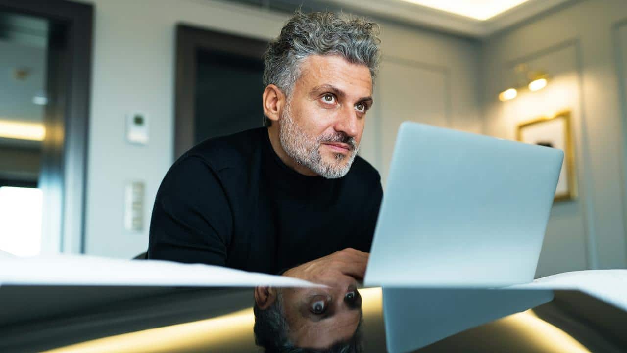 A man in a black turtleneck working on a laptop at a desk.