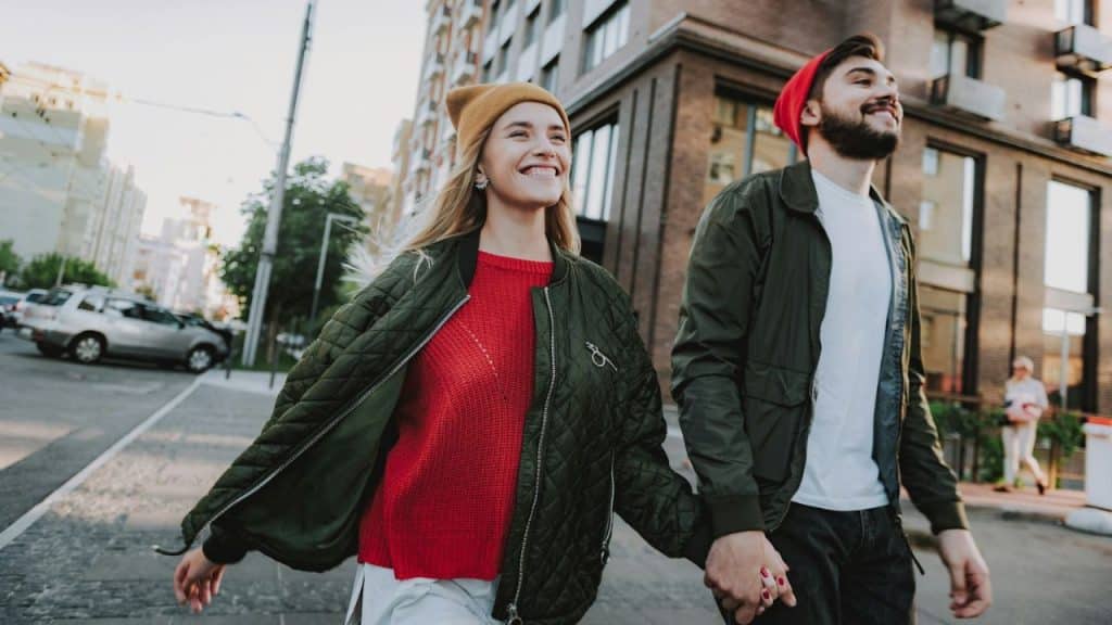 A smiling couple, wearing beanies and jackets, walks hand-in-hand outdoors.