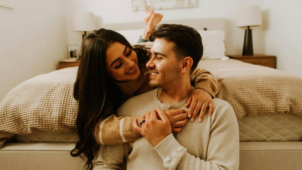 A couple sitting on the floor, smiling at each other in a cozy bedroom.