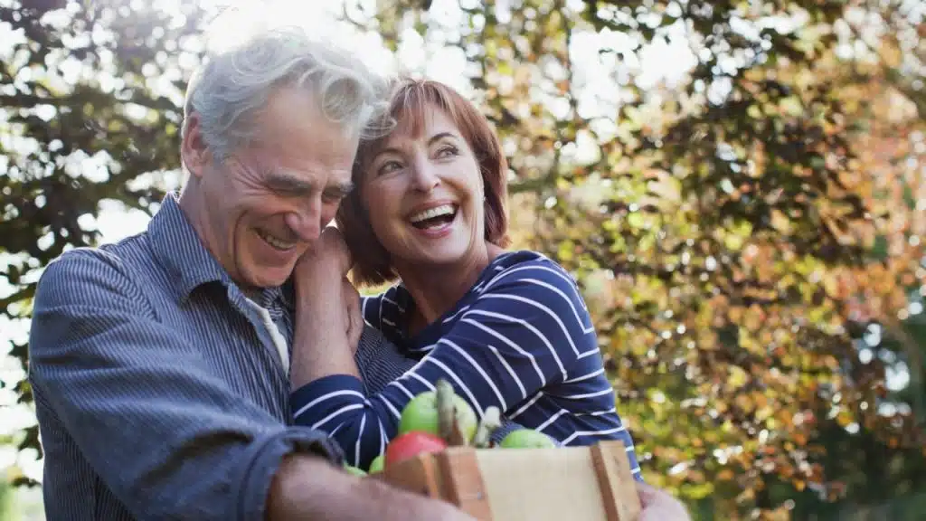A senior couple laughs outdoors, with the man holding a crate of apples and the woman embracing him.