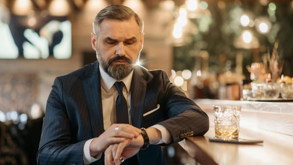 A businessman in a suit looks at his watch at a bar, a drink beside him.