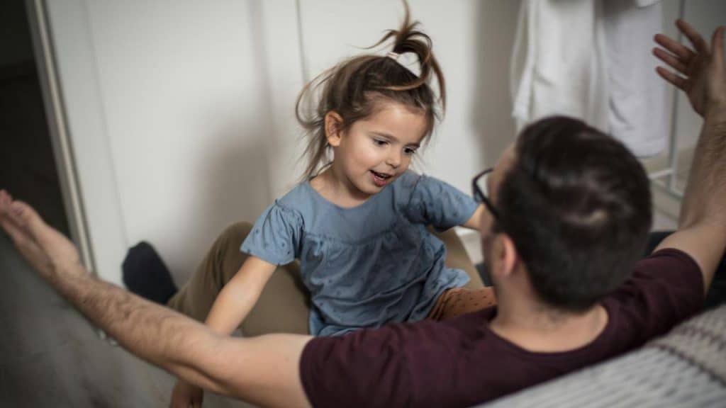 A man sitting on the floor playing with a smiling young girl in a casual indoor setting.