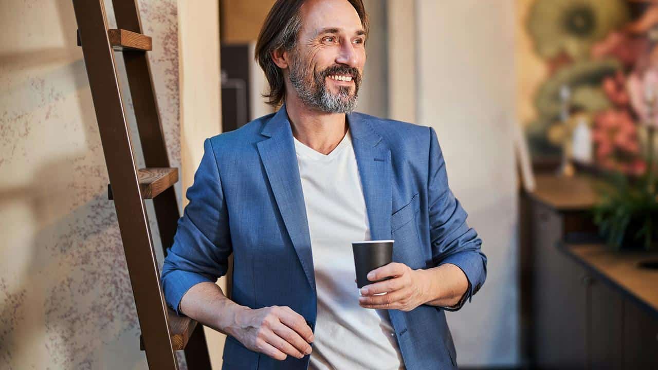 A man in a blue blazer holding a cup and leaning against a ladder.