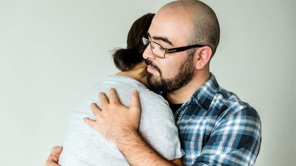 A man with a beard and glasses is hugging a person whose back is to the camera.