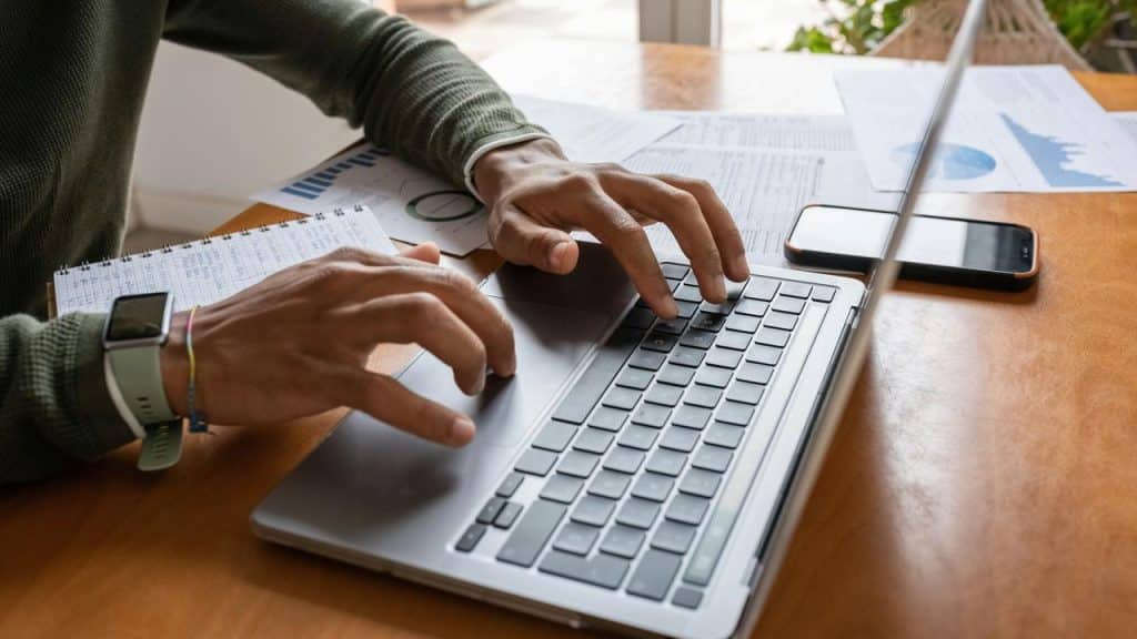 A person typing on a laptop at a desk with papers and a phone.