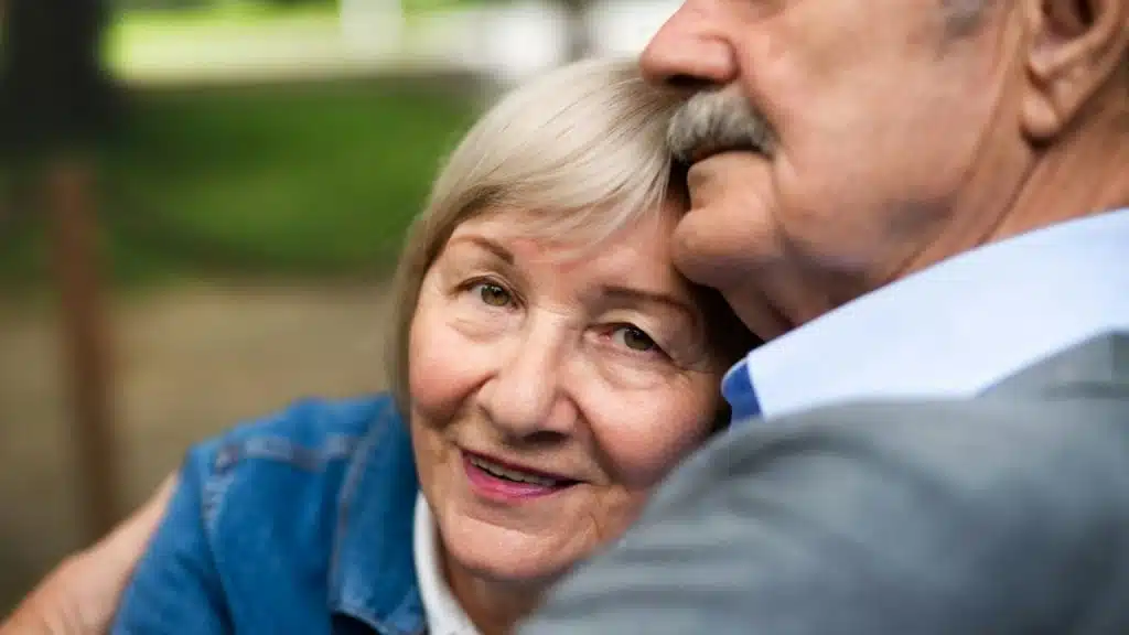 A senior woman with blonde hair, looking at the camera, is embraced by a man with a mustache.