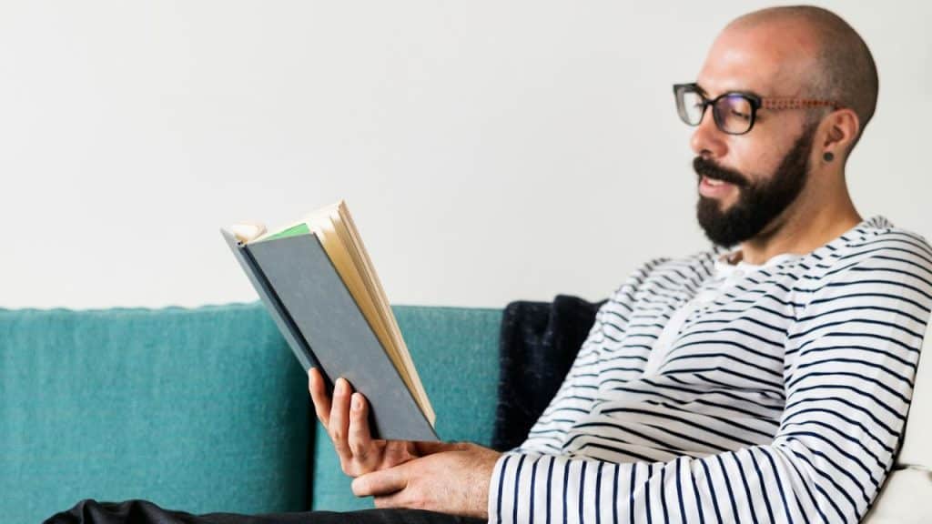 A bearded man with glasses, wearing a striped shirt, reads a book on a turquoise couch.