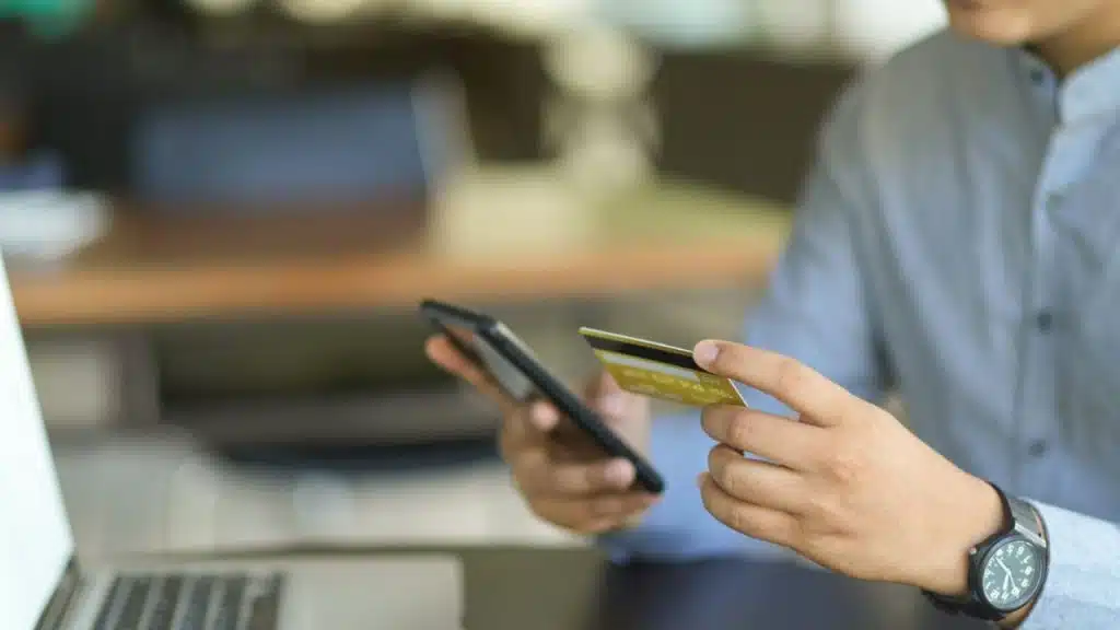 A person holds a credit card and a smartphone, with a laptop in the foreground.