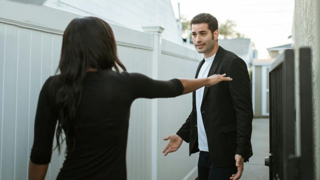 A woman in black extends her arm towards a man in a blazer looking confused.